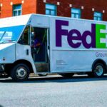 Delivery person in a green jacket standing beside a white and blue courier van, representing shipping services.